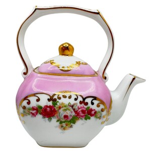 Puede incluir: Una tetera de porcelana blanca con una sección superior rosa, un asa con borde dorado y una tapa con pomo dorado. El cuerpo está decorado con un colorido patrón floral de rosas rojas y blancas, acentuado por intrincados adornos dorados.
