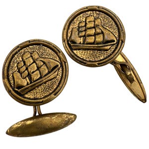 May include: A pair of vintage gold-toned cufflinks. Each cufflink features a detailed relief of a sailing ship within a circular frame. The cufflinks have an oval-shaped backing and a classic design.