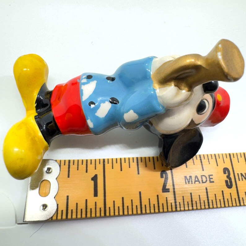 May include: A vintage Mickey Mouse figurine, approximately 6.3 cm long, in a playful pose with a gold-coloured horn. The figurine features a red shirt, blue jacket, yellow shoes, and black shorts. The figurine is made of ceramic.