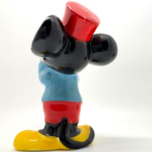 May include: A ceramic figurine of a cartoon character, with a black head and ears, a red hat, a light blue shirt, red shorts, and yellow shoes. The figurine has its arms crossed.