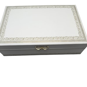 May include: A rectangular white storage box featuring a gold-colored filigree border pattern on the lid and a matching gold-colored metal latch on the front. The box has a smooth finish and a clean, structured design for holding various items.
