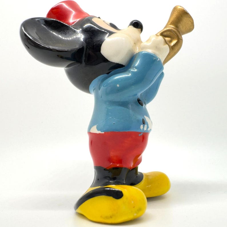 May include: A vintage Mickey Mouse figurine in a playing pose. The figure is painted in bright colours, including black, red, blue, and yellow. Mickey is wearing a red hat, blue jacket, and red shorts. He is holding a gold-coloured horn.