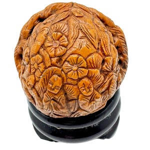 Może przedstawiać: Szczegółowa, ręcznie rzeźbiona drewniana rzeźba przedstawiająca kwiaty i postacie ludzkie. Rzeźba ma ciepły, jasnobrązowy kolor i jest zamontowana na czarnej, okrągłej podstawie. Złożone rzeźbienia pokazują umiejętności i dbałość o szczegóły artysty.