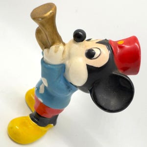 May include: A vintage Mickey Mouse figurine, with a red hat, blue jacket, yellow shoes, and a gold horn. The classic cartoon character is depicted in a playful pose, holding the horn. The figurine is made of ceramic and has a glossy finish.