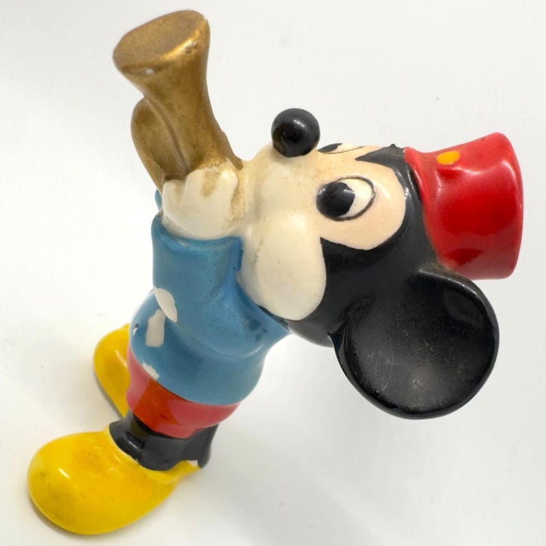 May include: A vintage Mickey Mouse figurine, with a red hat, blue jacket, yellow shoes, and a gold horn. The classic cartoon character is depicted in a playful pose, holding the horn. The figurine is made of ceramic and has a glossy finish.