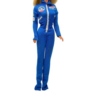 May include: A blue astronaut jumpsuit with a silver zipper and patches. The patches include a NASA logo and text. The jumpsuit has long sleeves and a fitted silhouette. The doll has blonde hair.