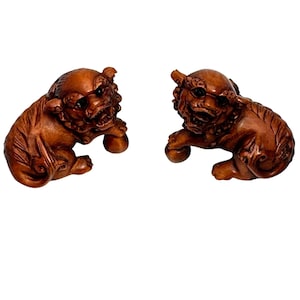 May include: Two carved wooden guardian lion statues in a reddish-brown hue. Each lion is depicted in a seated position, holding a small ball. The statues have detailed features, including expressive faces and textured manes, and are set against a white background.