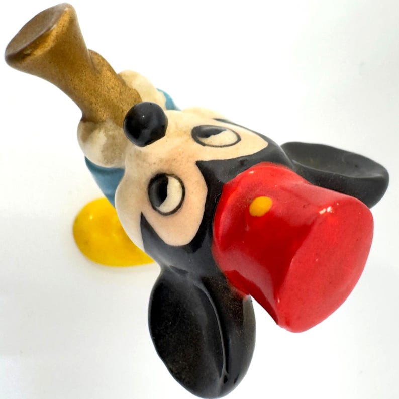 May include: A vintage Mickey Mouse figurine, with a red nose, black ears, and a cream-coloured face. The figure is holding a gold-coloured trumpet. The figurine has a yellow button and blue accents. A classic Disney collectible.
