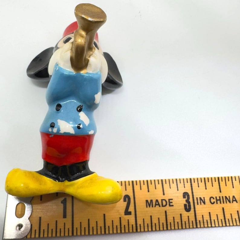 May include: A vintage Mickey Mouse figurine, painted in bright colours. Mickey is wearing a red hat, blue jacket, red trousers, and yellow shoes. He is holding a gold-coloured horn. The figurine is approximately 5 cm tall.