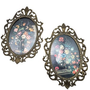 Puede incluir: Dos piezas de arte mural ovaladas vintage. Cada una presenta una impresión de naturaleza muerta floral sobre un fondo oscuro, enmarcada por metal dorado antiguo ornamentado. Los arreglos florales incluyen flores rosas, amarillas y azules.