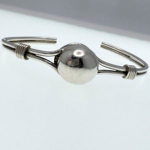 Op de afbeelding: Een zilverkleurige manchetarmband met een grote, bolvormige centrale kraal. De armband heeft een minimalistisch ontwerp met draaddetails en een gepolijste afwerking. Het open ontwerp maakt het gemakkelijk om te dragen.