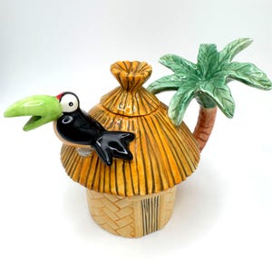 Puede incluir: Una tetera de cerámica con forma de choza de techo de paja y base beige texturizada. Un tucán negro con pico verde brillante está posado en el techo. Una palmera verde se extiende desde el lateral como asa, con una tapa a juego en forma de choza.