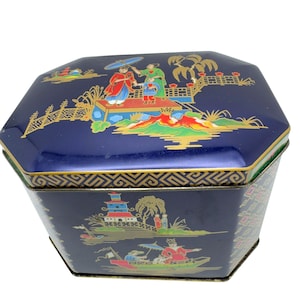 Può includere: Una scatola di metallo ottagonale blu scuro con illustrazioni dorate, rosse e verdi di figure, pagode e barche. Il coperchio mostra figure sotto un ombrello vicino a un ponte, mentre i lati presentano scene paesaggistiche con edifici.