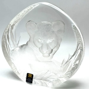 May include: A clear, circular glass sculpture featuring a frosted, three-dimensional relief of a lion cub. The piece has a smooth, polished outer edge and a textured, jagged border. A black label at the bottom reads Mats Jonasson Sweden.
