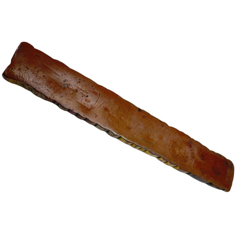 May include: A long, rectangular wooden object in reddish-brown. The surface is textured with markings, and a decorative edge is visible. It appears to be a decorative item or part of a larger piece. The object is approximately 30 cm long.
