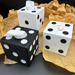 May include: Three tissue box covers shaped like dice. One is black with white dots, one is white with black dots, and one is white with black dots. The tissue boxes are sitting on brown crumpled paper.