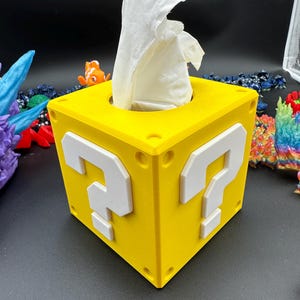 May include: A yellow question mark block tissue box holder with white question marks on each side. The tissue box is made of plastic and has a hole in the top for tissues.