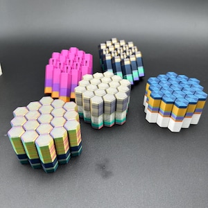 May include: Five hexagonal shaped coasters made of stacked, colorful, hexagonal tiles. The coasters are in various colors, including pink, gray, blue, and iridescent.