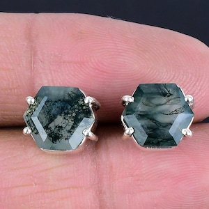 May include: A pair of hexagonal stud earrings. Each earring features a green and white moss agate stone set in a silver metal setting. The earrings are displayed against a dark background.