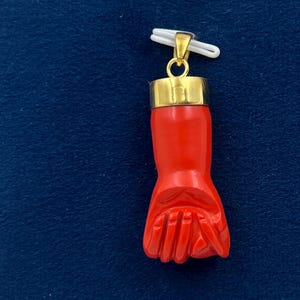 Puede incluir: Un colgante de mano de coral rojo con parte superior dorada. La mano está tallada en forma de puño con los dedos cerrados.