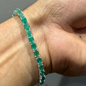 Op de afbeelding: Een zilveren armband met een rij ovaal geslepen smaragden. De armband wordt om een pols gedragen, waarbij de levendige groene edelstenen worden getoond. De armband is een sieraad.