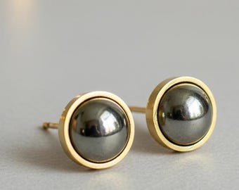 Op de afbeelding: Een paar oorstekers met een donkergrijze, bolvormige steen gezet in een goudkleurige bezel. De oorbellen worden gepresenteerd op een lichtgrijze ondergrond, met zachte, onscherpe lichten op de achtergrond. Klassieke sieraden.
