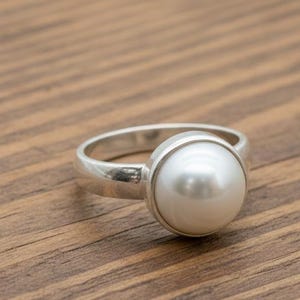 May include: A silver ring featuring a large, round, white pearl. The pearl is set in a bezel setting, and the ring band is smooth and polished. The ring is displayed on a wooden surface.