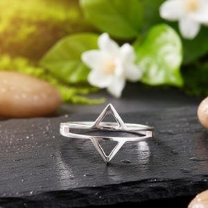 May include: A silver ring with a geometric star design. The ring features two connected triangles, creating a star shape. The ring is displayed on a dark, textured surface with wet stones and greenery in the background.