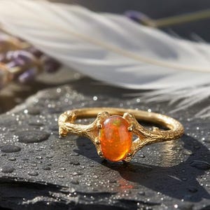 Puede incluir: Un anillo de oro con una piedra preciosa ovalada de color naranja. El anillo tiene una banda texturizada con un diseño similar a una rama. Se encuentra sobre una superficie oscura y húmeda, con una pluma blanca y ramitas de lavanda en el fondo.