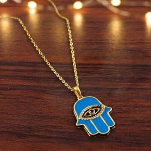 May include: A gold-colored necklace with a blue Hamsa hand pendant. The pendant features an eye design with clear crystals and a black center. The necklace is displayed on a wooden surface with string lights in the background.