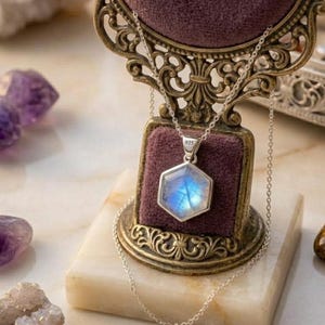 May include: A silver necklace featuring a hexagonal pendant with a translucent moonstone displaying a blue iridescent sheen. The pendant is stamped with 925 and hangs from a delicate silver chain, displayed on a dark purple velvet stand.