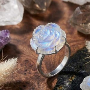 May include: A silver ring featuring a carved moonstone rose with a translucent, iridescent blue glow. The gemstone is set in a four-prong metal mounting, showcasing the intricate petal details of the floral carving against a polished band.
