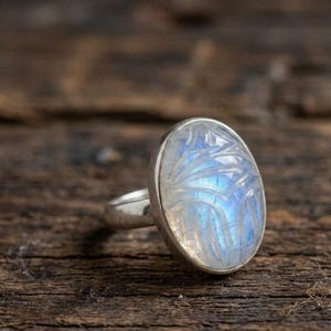 May include: A silver ring featuring an oval, carved moonstone. The moonstone has a blue and white iridescent glow, with a detailed, etched design. The ring is set on a dark wooden surface.