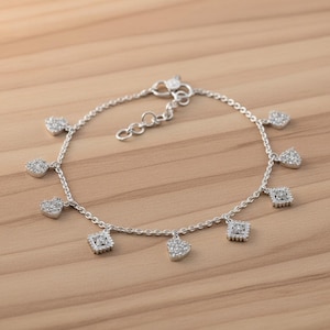 Puede incluir: Una pulsera plateada con múltiples colgantes en forma de corazón y cuadrado. Cada colgante está incrustado con pequeñas piedras transparentes. La pulsera se muestra sobre una superficie de madera marrón claro.