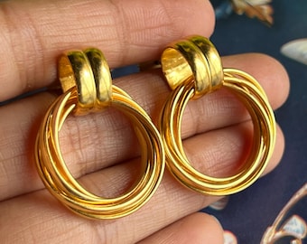以下が含まれることがあります： 金色のイヤリングのペア。各イヤリングは、一連の絡み合った金色のリングに接続された、厚く丸みを帯びた上部を備えています。イヤリングはぼやけた背景に対して表示され、金属的な輝きと複雑なデザインを強調しています。
