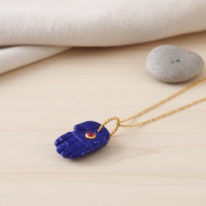 May include: A gold chain necklace featuring a blue hand-shaped pendant. The pendant has a small red gemstone. The necklace rests on a light wooden surface, with a grey stone and white cloth in the background.