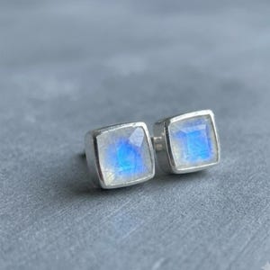 May include: A pair of square stud earrings featuring faceted moonstones set in silver metal frames. Each stone displays a translucent white appearance with a distinct blue adularescence glow emanating from the center of the square cut surface.