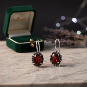 May include: Silver drop earrings featuring oval, deep red gemstones, each framed by a beaded edge. The earrings are presented on a marble surface, with a green velvet jewellery box visible in the background.