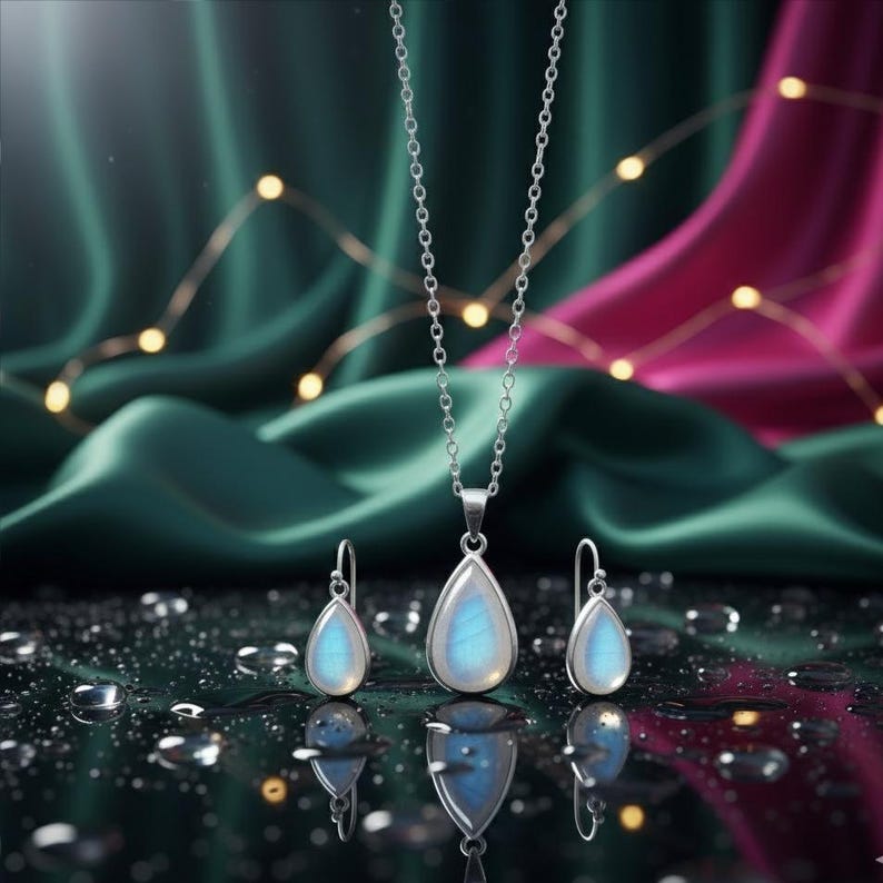 May include: A silver necklace and matching earrings set. The pendant and earrings feature a pear-shaped, iridescent stone. The jewellery is displayed on a reflective surface with water droplets, against a backdrop of green and pink fabric.
