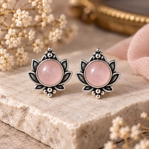 Puede incluir: Un par de pendientes de botón con una piedra preciosa redonda, lisa y de color rosa pálido, engastada en una montura de plata oxidada oscura con forma de flor de loto, con pétalos puntiagudos y pequeñas esferas decorativas en los extremos.
