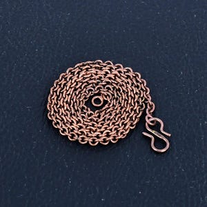 Puede incluir: Un collar de cadena de cobre con cierre de mosquetón. La cadena está enrollada en un círculo y el cierre cuelga hacia un lado.