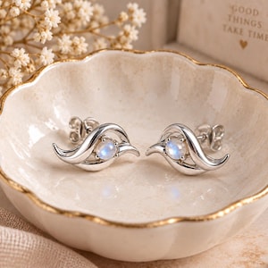 May include: A pair of silver stud earrings featuring a polished, curved metal design that wraps around a central, round, iridescent moonstone. The earrings have a secure post back closure and exhibit a smooth, reflective finish on the metal surface.