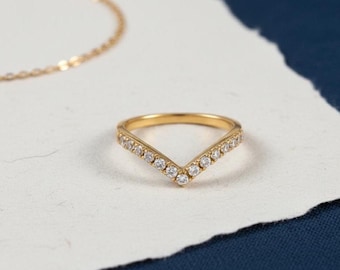May include: A gold-colored V-shaped ring with small, clear gemstones set along the top edges. The ring sits on a white surface, with a gold necklace chain visible in the background. The background is a dark blue color.