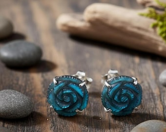 May include: A pair of teal rose-shaped stud earrings with silver-colored settings. The earrings are carved with detailed petals and are displayed on a wooden surface with smooth stones and driftwood in the background. The earrings are a jewelry item.