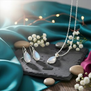 May include: A silver necklace and matching earrings set with teardrop-shaped moonstone pendants. The jewellery is displayed on a black slate surface with white flowers and a teal satin backdrop. The moonstones have a pearlescent sheen.