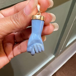 Könnte beinhalten: Hellblauer Handanhänger mit goldfarbener Kappe. Der Anhänger ist in Form einer Hand mit detaillierten Fingern geschnitzt. Die goldene Kappe hat eine Schlaufe für eine Kette. Der Anhänger wird vor einem neutralen Hintergrund gehalten.
