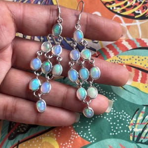 Könnte beinhalten: Hängende Chandelier-Ohrringe mit ovalen Opalen in Silber gefasst. Die Opale zeigen eine Reihe von Farben, darunter Blau, Grün und Weiß. Die Ohrringe werden von einer Hand gehalten.