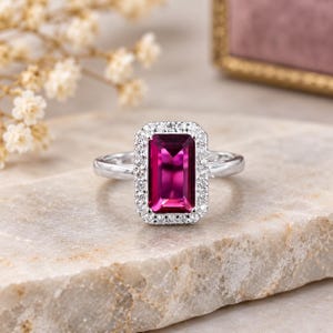 May include: A silver-toned ring featuring a large, rectangular, deep pink gemstone set in the center. The central stone is surrounded by a halo of small, clear, round-cut stones. The band is smooth and metallic, extending from the central setting.