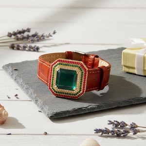 May include: A brown leather watch with a large, emerald-cut green gemstone. The watch face is surrounded by red and green gemstones and a gold frame. The watch band is brown with a crocodile texture.