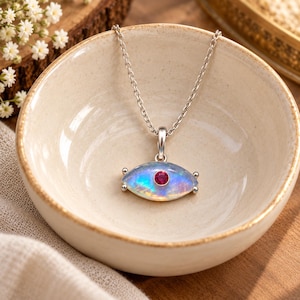 May include: A silver necklace featuring an eye-shaped pendant with an iridescent opal-like stone and a small, round, vibrant pink gemstone set in the center. The pendant hangs from a delicate silver chain, resting inside a small, cream-colored bowl.